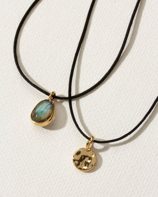 Two black corded necklaces, one with a labradorite charm, and the other with a gold hammered charm, by Luna Norte Jewelry.