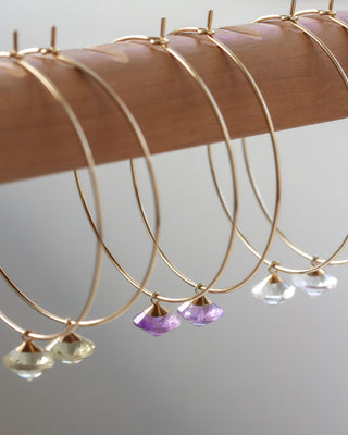 Several pairs gold hoop earrings with gemstone drops on a wooden hanger against a neutral background