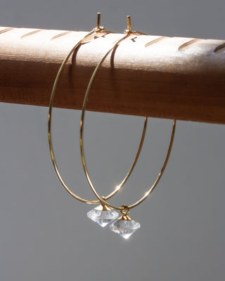 Gold hoop earrings with clear quartz gemstones on a wooden stick against a gray background