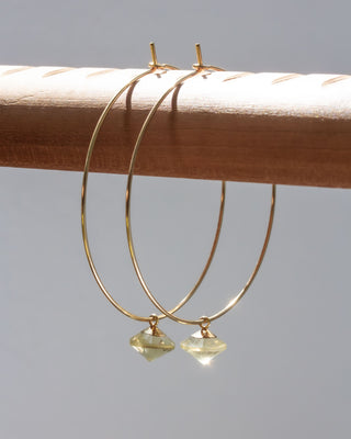 Gold hoop earrings with greenish gemstones on a wooden dowel against a gray background