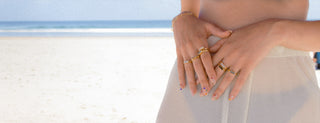 Close-up of hands with various gemstone gold rings on a blurred beach background