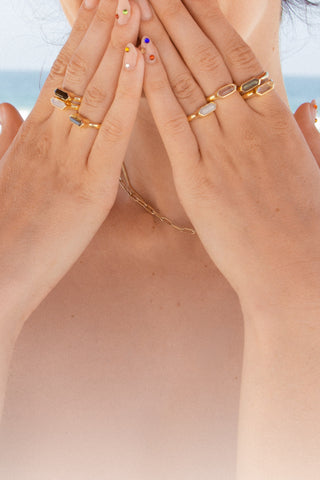 A close-up lifestyle shot of a person's hands covering their face, showcasing a collection of gold gemstone rings and bedazled press on nails