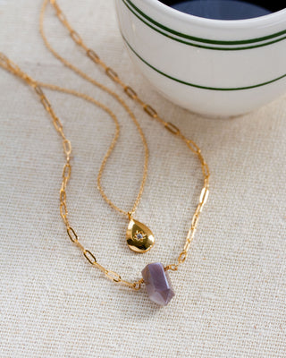 Two gold-plated necklaces, one with a faceted coffee moonstone gemstone pendant and the other with a golden teardrop charm, laid out on a textured surface next to a white ceramic cup with green accents.