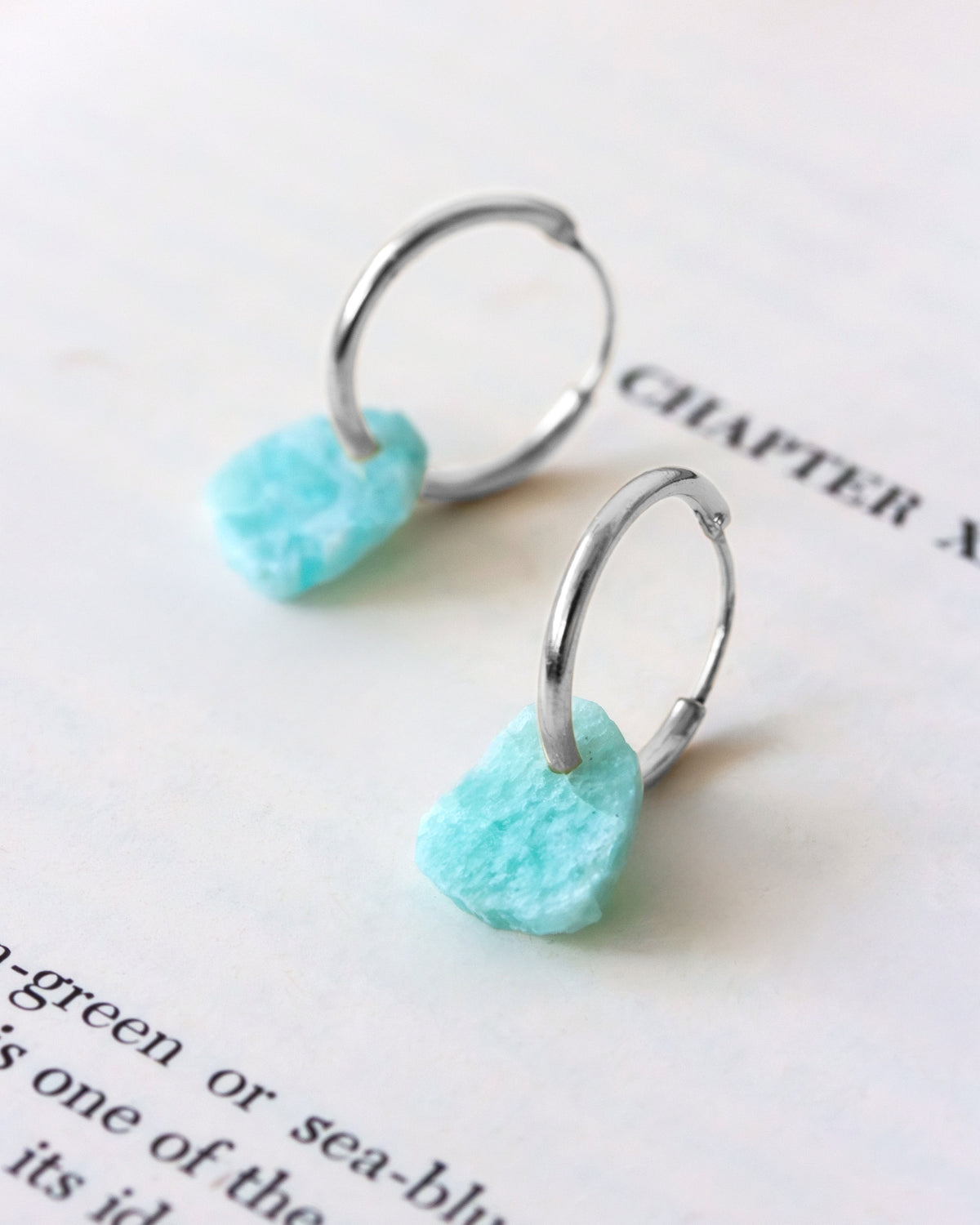 A pair of silver-plated hoop earrings with raw-edge amazonite gemstones on book page.