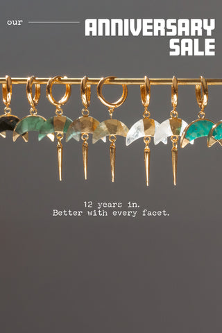 Various Gold gemstone hoop earrings hanging on a gold bar in front of a gray background with text overlay that reads "Our Anniversary Sale, 12 Years in. Better with every facet"