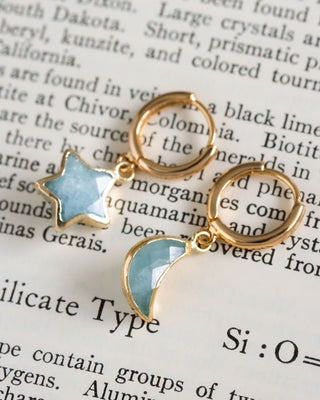 A pair of mismatched gold huggie hoop earrings with aquamarine star and moon charms, set against a background of text.