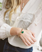 A close-up of a woman wearing a gold chain bracelet with a green heart charm, styled with a white lace blouse and dark blue jeans.