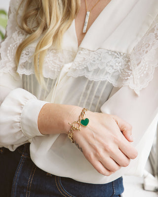 A close-up of a woman wearing a gold chain bracelet with a green heart charm, styled with a white lace blouse and dark blue jeans.