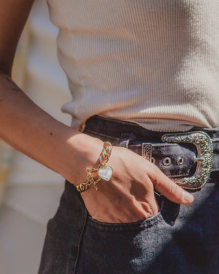 A close-up lifestyle shot of a person wearing a gold chain bracelet with a moonstone heart-shaped charm, styled with a dark brown textured belt and dark denim jeans.