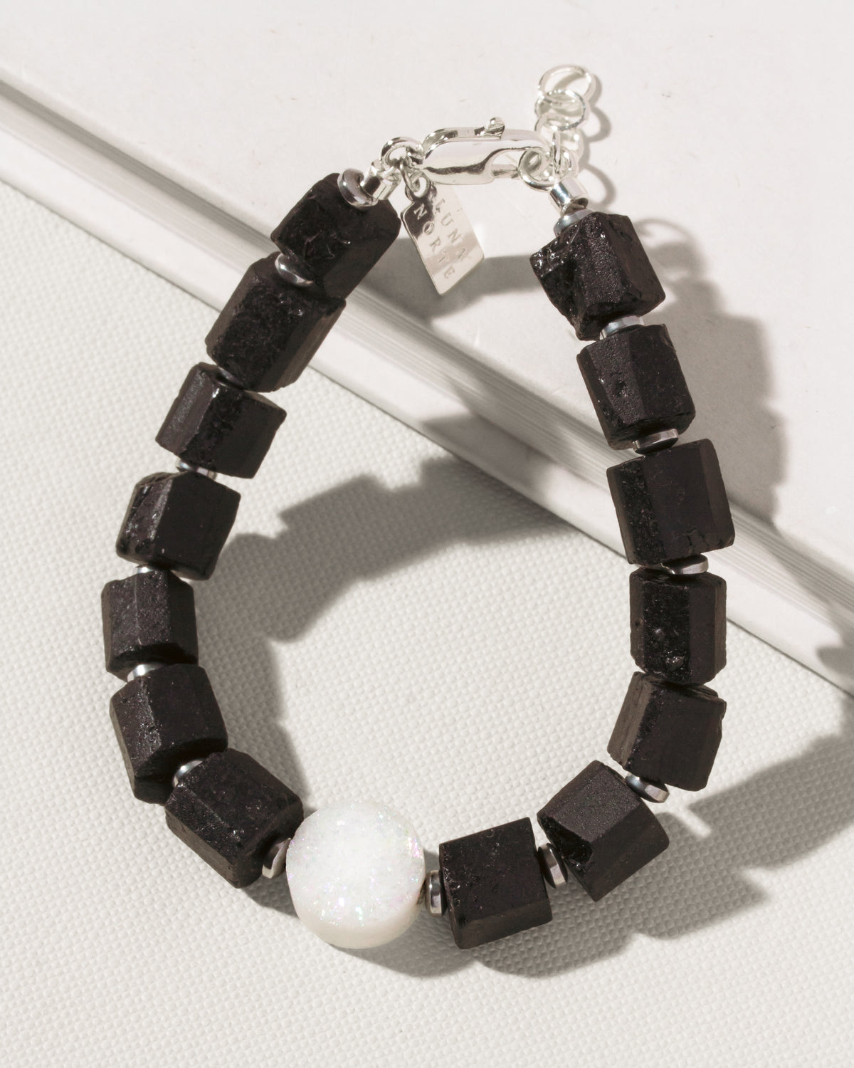 A beaded  bracelet featuring matte black cube-shaped tourmaline beads, a single white round druzy bead, and silver-toned metal spacers and clasp, displayed on a textured light gray surface.