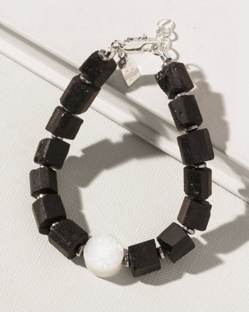 A beaded  bracelet featuring matte black cube-shaped tourmaline beads, a single white round druzy bead, and silver-toned metal spacers and clasp, displayed on a textured light gray surface.