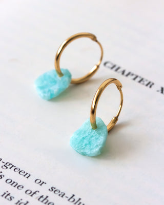 A pair of gold-plated hoop earrings with raw-edge amazonite gemstones attached, displayed on a piece of paper.