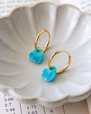 A pair of gold-plated hoop earrings with raw-edge turquoise gemstones attached, displayed in a ceramic floral bowl.
