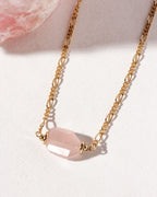 A gold-colored  necklace with a rose quartz faceted pendant, displayed on a textured background.