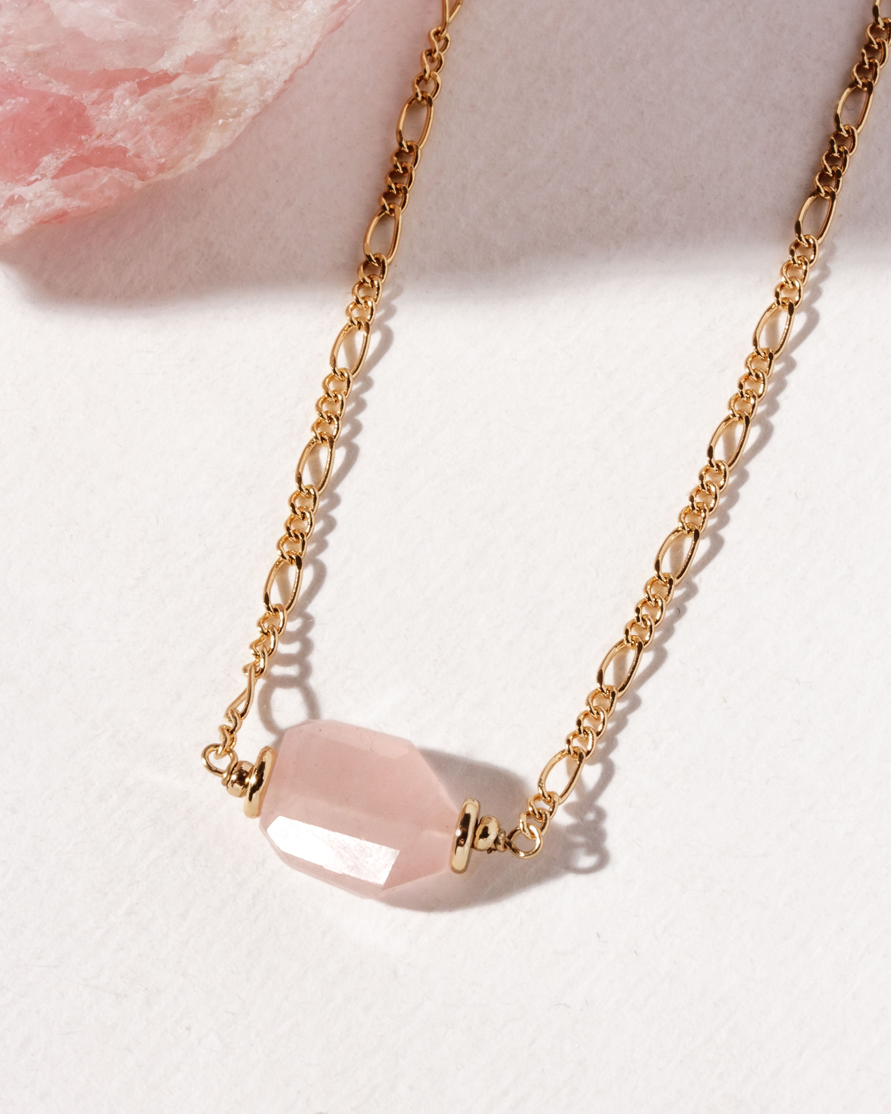 A gold-colored  necklace with a rose quartz faceted pendant, displayed on a textured background.