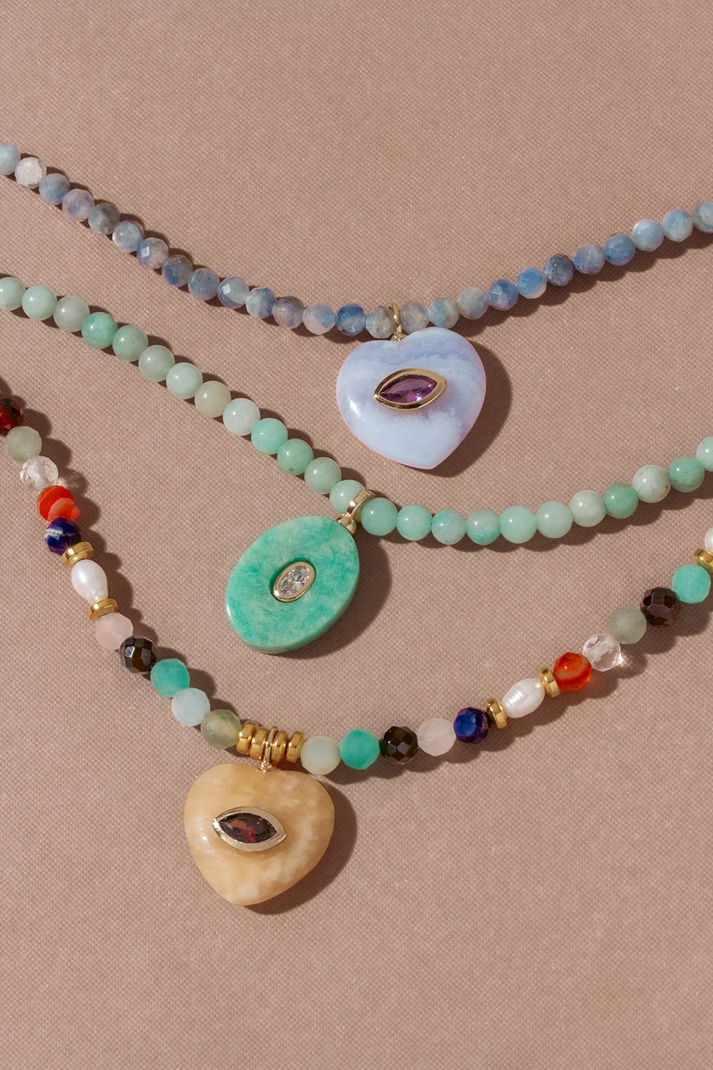 Three layered beaded necklaces with evil eye charms inlaid into genuine gemstone pendant, displayed on a textured beige background. The necklaces feature various beads in green, blue, white, brown, and red, with gold-toned metal accents.