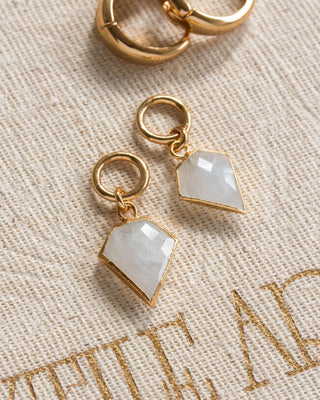 A pair of gold-colored interchangeable hoop earring charms with a moonstone, resembling a diamond, placed on a beige fabric surface.