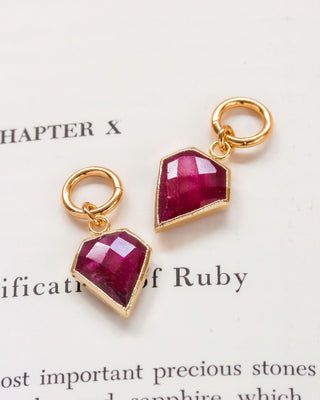 A pair of gold-colored interchangeable hoop earring charms with a faceted ruby gemstone, resembling a diamond, placed on a book page.