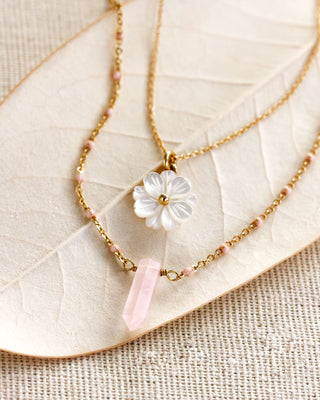 A gold layered necklace set with a carved mother of pearl shell daisy and a pink rose quartz crystal pendant, displayed on a fabric surface.