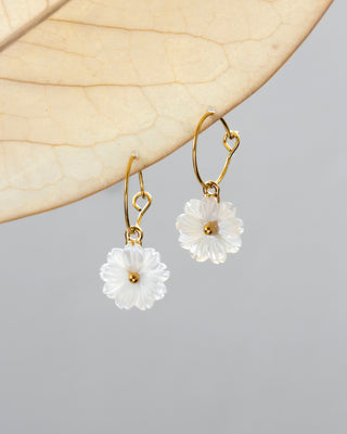 A pair of gold-colored hoop earrings with white mother of pearl shell daisy designs attached to the hoops, displayed against a beige background.