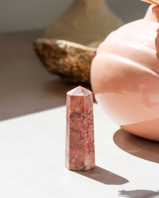 A mini terra cotta calcite tower with a smooth, faceted shape, placed on a flat surface, with a blurred background featuring a textured object.