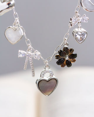 A silver charm necklace featuring various shapes including hearts, a moon, a star, and a flower, with polished silver hearts, CZ pavé bow pendant, and a black lip mother of pearl flower.