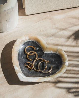 A heart-shaped, black and yellow two-toned Calcite jewelry catchall dish with a smooth polish finish, containing golden earrings inside, placed on a wooden surface with a shadow cast over it.
