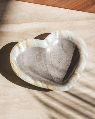 A heart-shaped, grayish two-toned calcite, jewelry catchall dish with a smooth polish finish, placed on a wooden surface with a shadow cast over it.