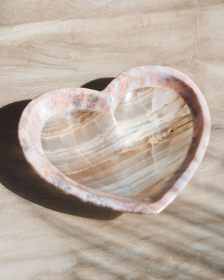 A heart-shaped, pinkish two-toned calcite jewelry catchall dish with a smooth polish finish, placed on a wooden surface with a shadow cast over it.