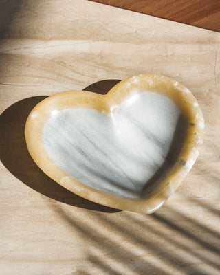 A heart-shaped, two-toned Calcite jewelry catchall dish with a smooth polish finish, placed on a wooden surface with a shadow cast over it.