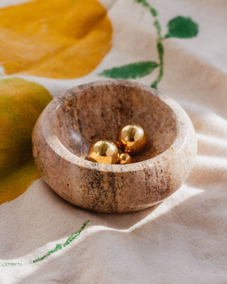 A small round calcite catchall bowl with a smooth finish, containing a few golden jewelry items, placed on a cloth with leaf patterns.