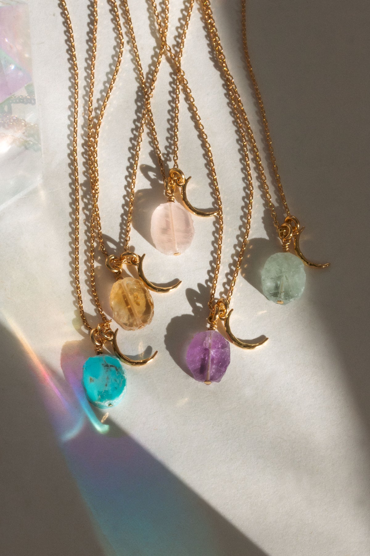 Five gold crescent moon and chain necklaces with freeform oval gemstone pendants in turquoise, amethyst, aquamarine, citrine, and rose quartz, arranged on a white surface with dramatic sunlight and shadow.