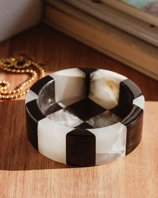 A round jewelry catchall dish with a black and white checkerboard pattern, made from genuine stone, placed on a wooden surface.