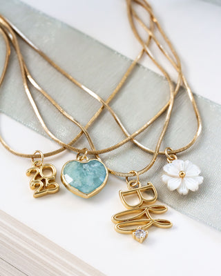 Gold layered necklaces with 2025, BFF, aquamarine heart, and mother-of-pearl floral charms on a light background