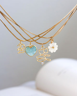 Layered gold necklaces with 2025, BFF, Aquamarine heart, and mother-of-pearl floral charms on a light gray background.