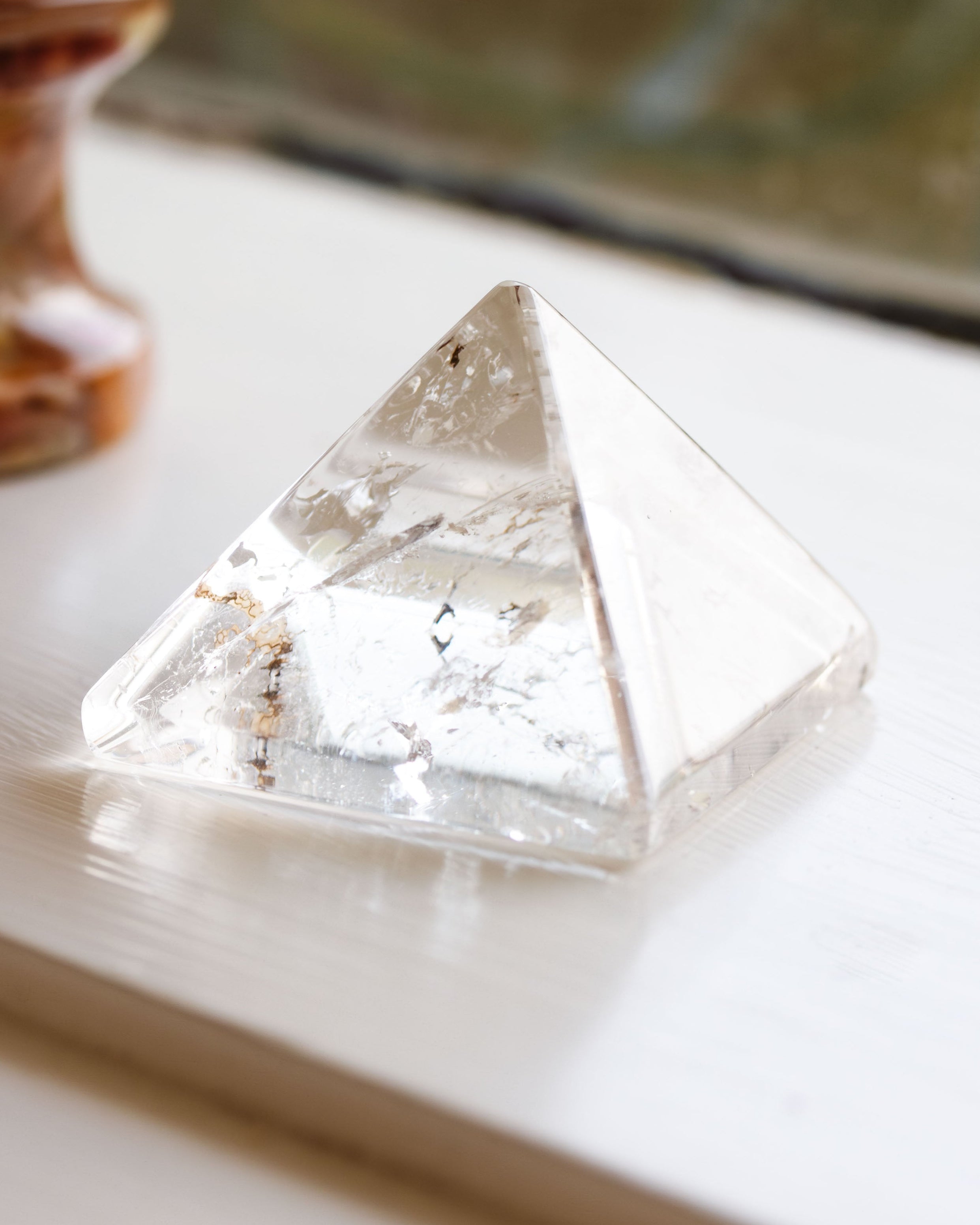 A clear crystal quartz pyramid placed on a wooden surface, with a blurred object in the background.