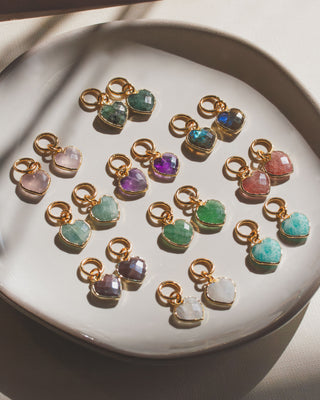 Off white dish containing many different  gemstone pairs of heart shaped gold hoop earrings.