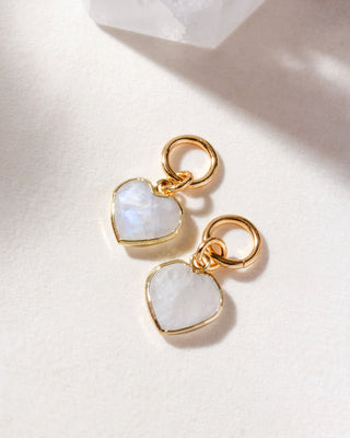 A pair of interchangeable hoop earring charms with heart shaped moonstone gemstones displayed on a white surface.