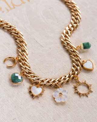 A chunky gold chain charm necklace with rose quartz, emerald, mother of pearl, and druzy moon, floral, heart, sun, and pendant charms on a beige fabric background.