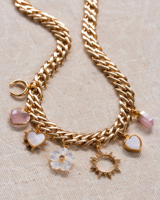 A chunky gold chain charm necklace with rose quartz, mother of pearl, and druzy charms on a beige fabric background.