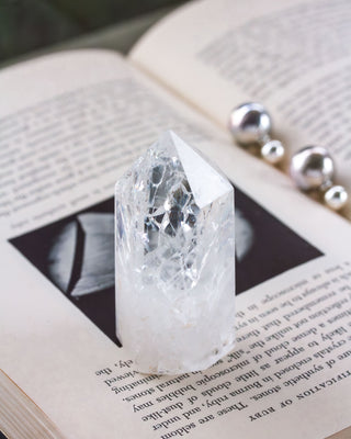 A clear quartz gemstone tower with a crystalline appearance, placed on an open book, with a black and white image in the background and spherical objects to the side.