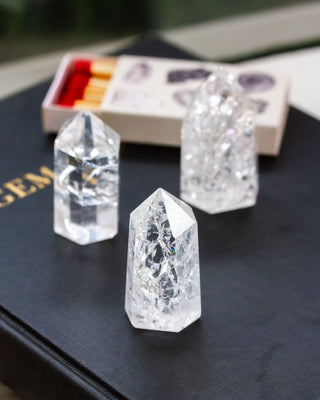 Three clear quartz crystal towers of varying sizes, displaying a crackle effect, placed on a dark surface with a blurred box of matches in the background.