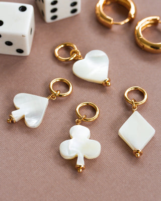 Four interchangeable hoop earring charms made of mother of pearl, carved into hearts, clubs, spades, and diamonds shapes, displayed on a surface with dice and gold-colored earrings in the background.