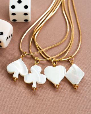 A collection of gold snake chain necklaces with carved mother-of-pearl card suit symbols pendants, displayed against a beige background with dice for scale.