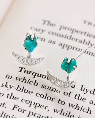 Pair of silver turquoise post earrings with crescent moon and cz embeddings, on a book page.