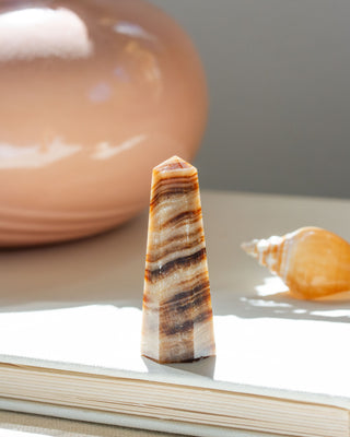 A small calcite mini obelisk with desert stripes, placed on a flat surface, with a blurred background featuring a reflective surface and a shell.