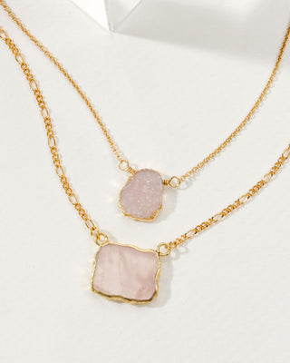 A layered necklace with a gold chain and pink Druzy and rose quartz pendants displayed on a white surface.