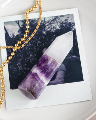 A faceted crystal tower with clear quartz on top and deep amethyst on the bottom, displayed on a white surface with a gold chain and a polaroid in the background.