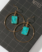 A pair of gold hoop earrings with rectangular turquoise-blue stone pendants, displayed on a black notebook with a grid pattern.
