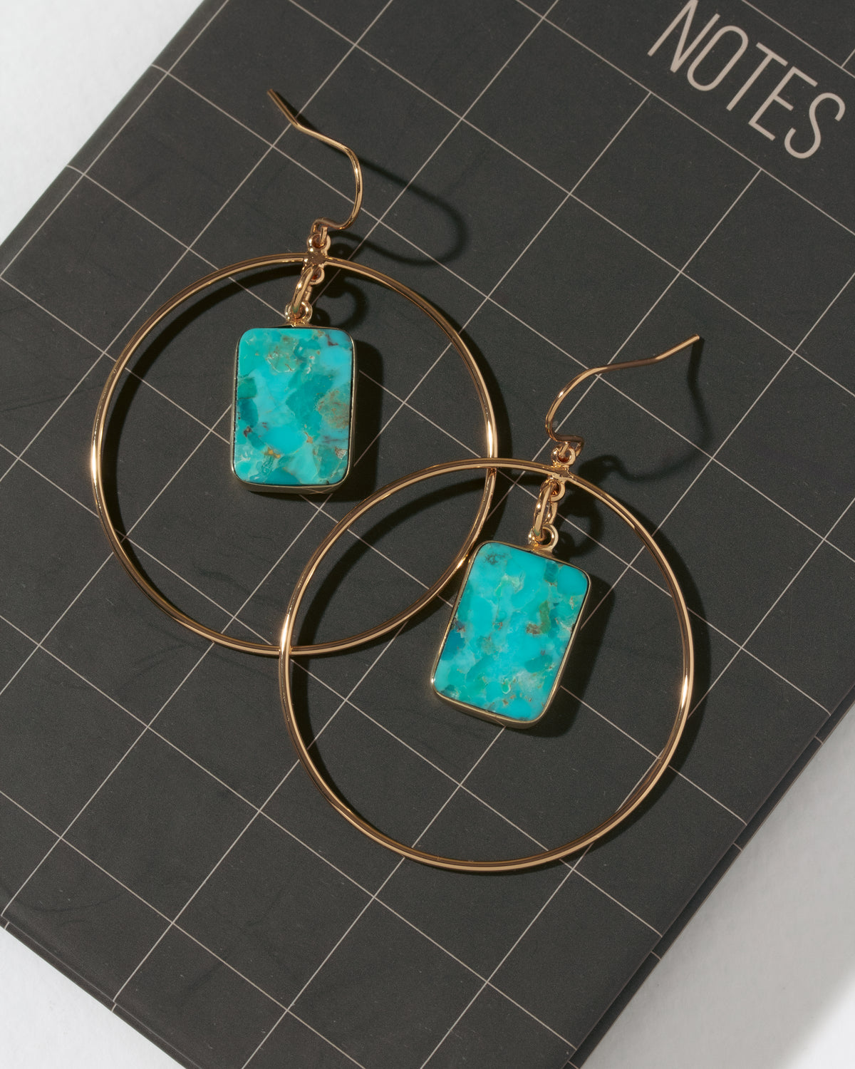 A pair of gold hoop earrings with rectangular turquoise-blue stone pendants, displayed on a black notebook with a grid pattern.